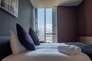 Un cuarto de hotel con un ventanal grande, una cama con ropa blanca y vistas a la montaña.