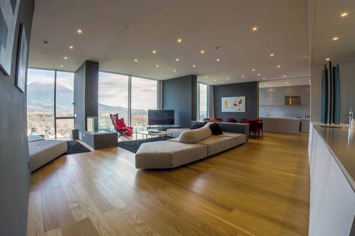 A spacious living area with a large window, a modern sofa, a red chair, and a kitchen in the background.