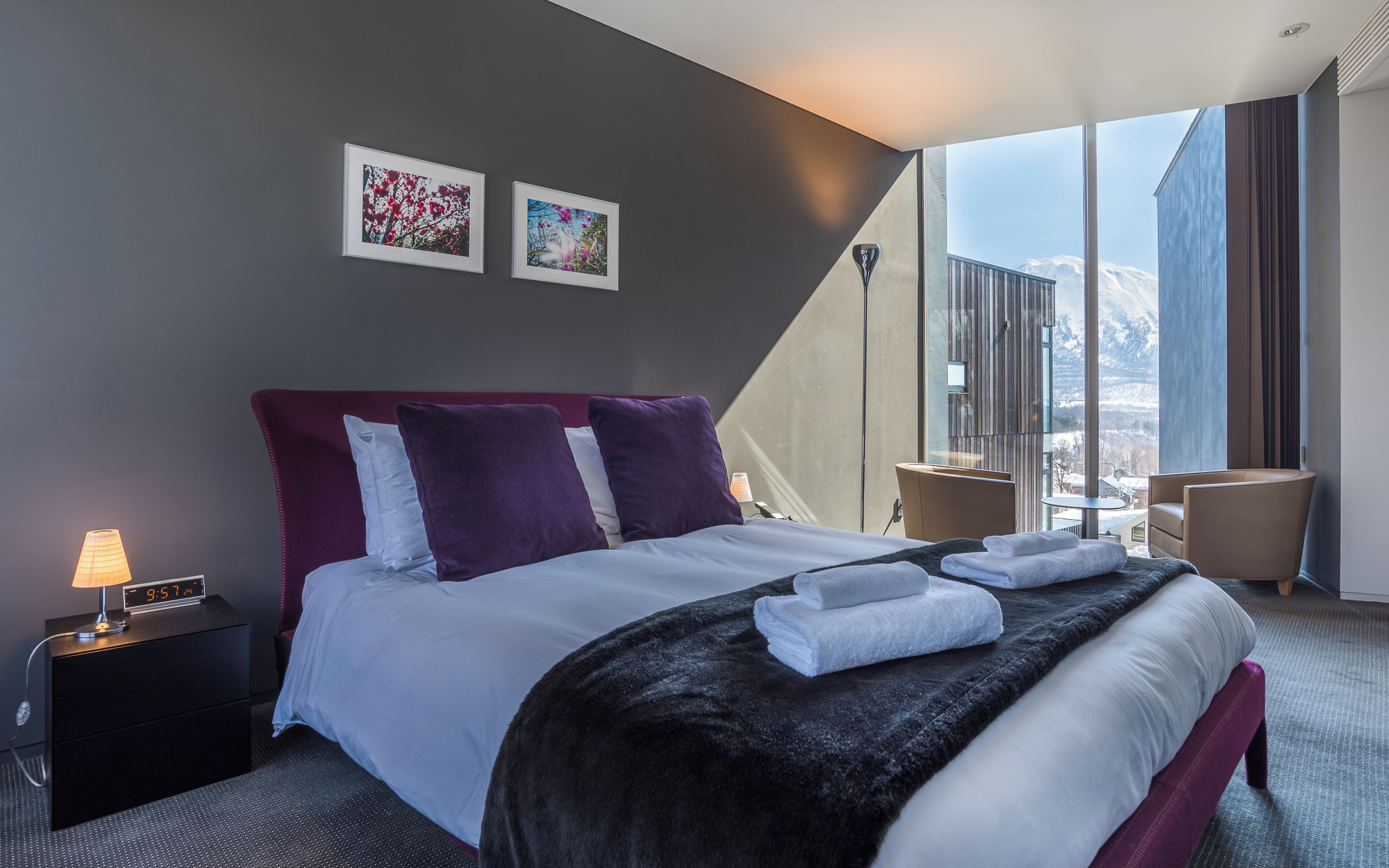 A modern hotel room with a large bed, a nightstand with a lamp, two framed pictures on the wall, and a view of mountains through large windows.