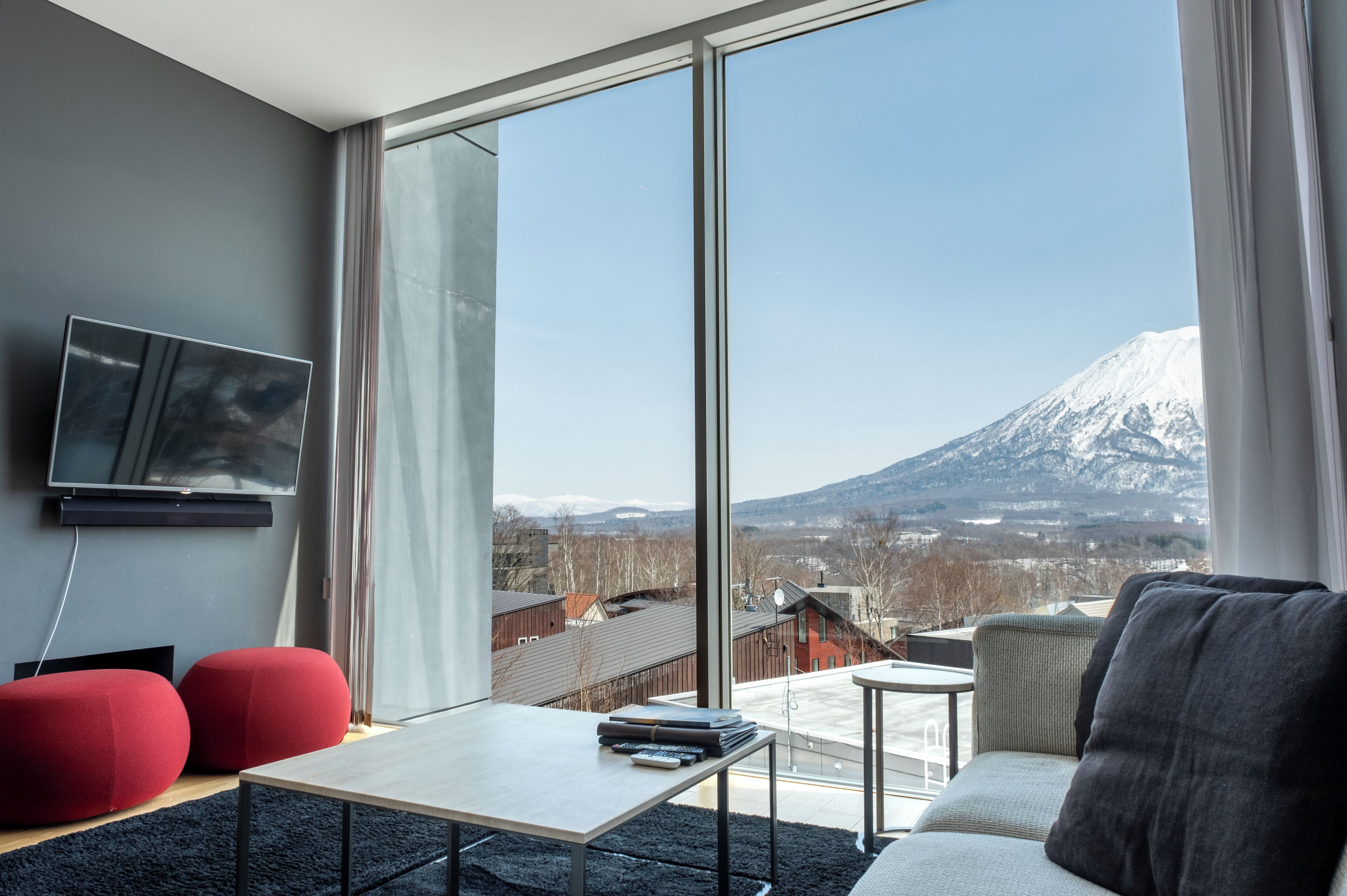 Un salon moderne doté d’une grande fenêtre, d’un téléviseur à écran plat, de poufs rouges et offrant une vue sur une montagne enneigée.