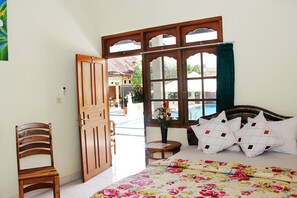 A bedroom with a bed, a wooden chair, a window with a view of a pool and buildings, and a small table with a vase of flowers. - Warung Coco Hostel (Kuta)