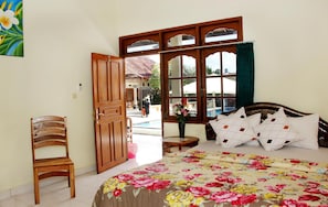 A bedroom with a bed, a wooden chair, a window with a view of the outside, and a door leading to another room. - Warung Coco Hostel (Kuta)