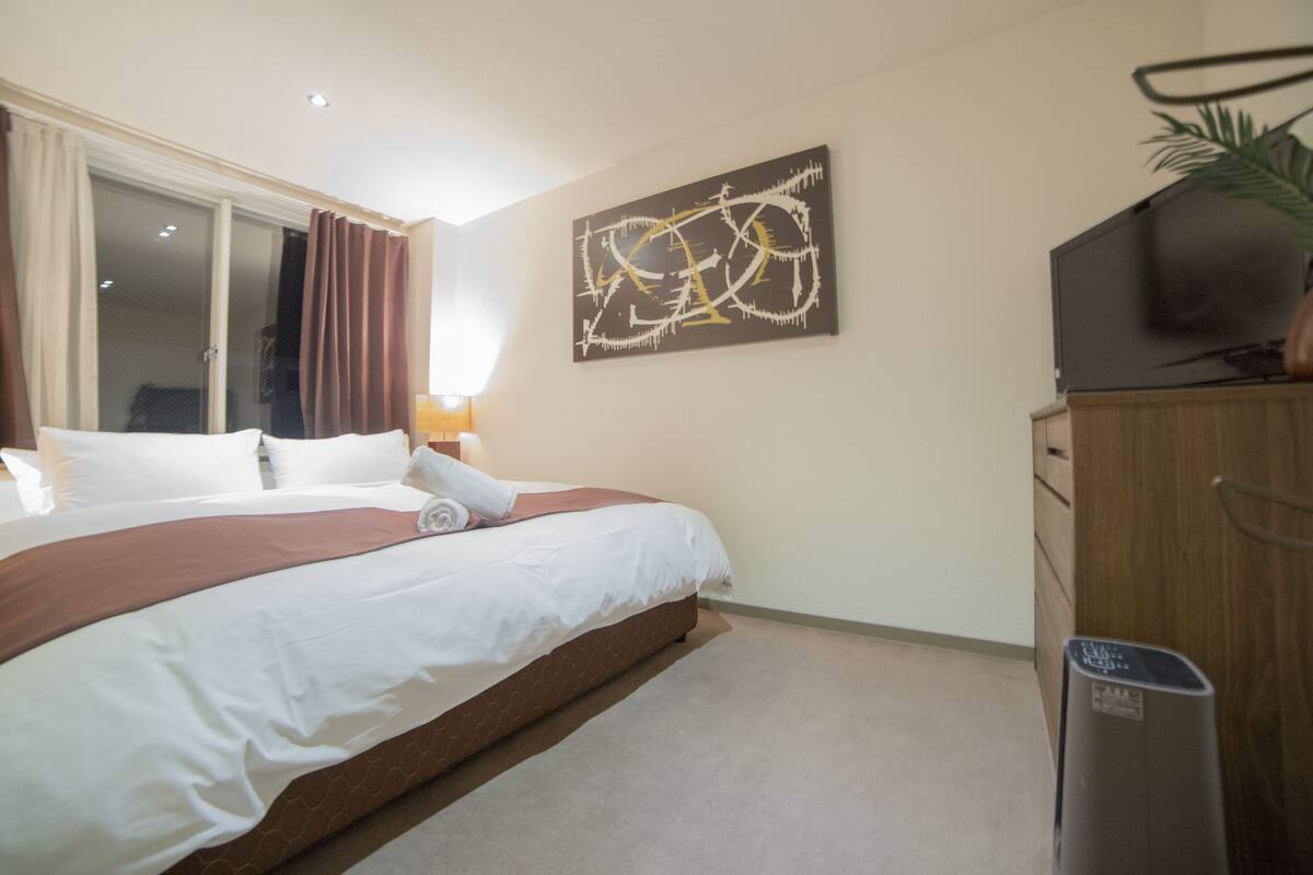 A hotel room with a bed, a television, a waste bin, and a painting on the wall.