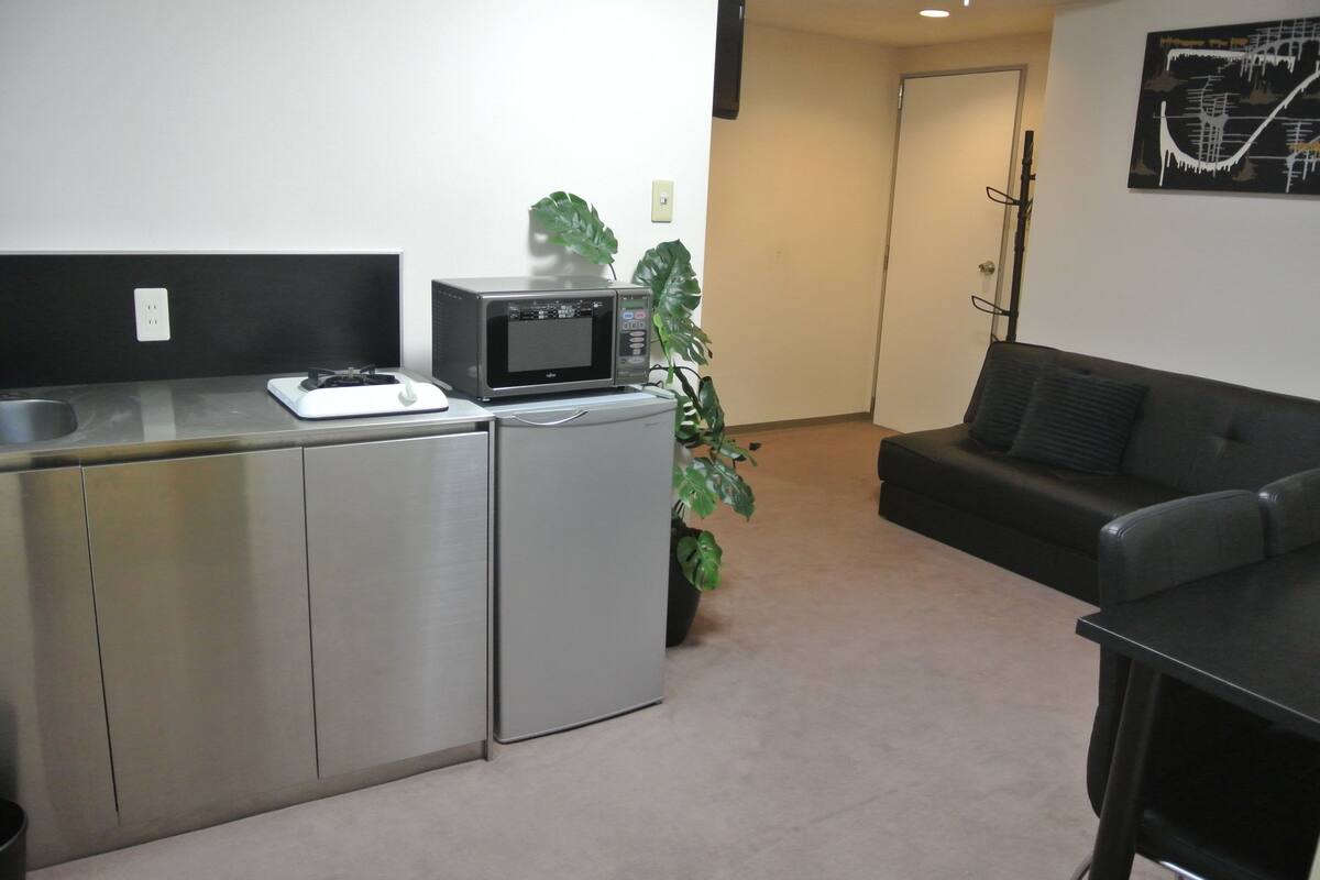 A compact kitchenette with a microwave, a small refrigerator, and a sink. There is a potted plant and a black couch in the room.