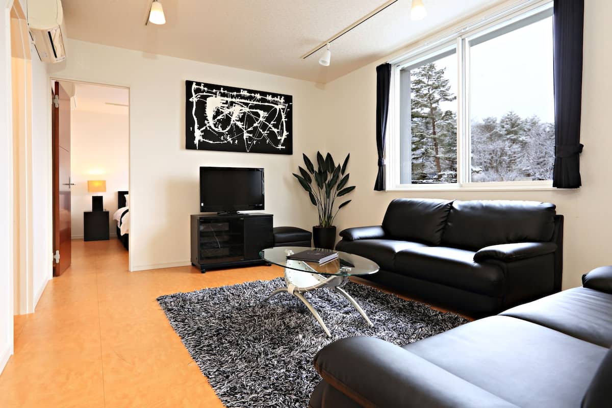 A modern living room with a black leather sofa, a glass coffee table, and a large window with a view of snow-covered trees.
