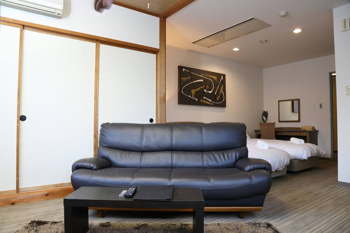A modern hotel room with a leather sofa, a small table, a bed with white linens, a painting on the wall, and a sliding door.