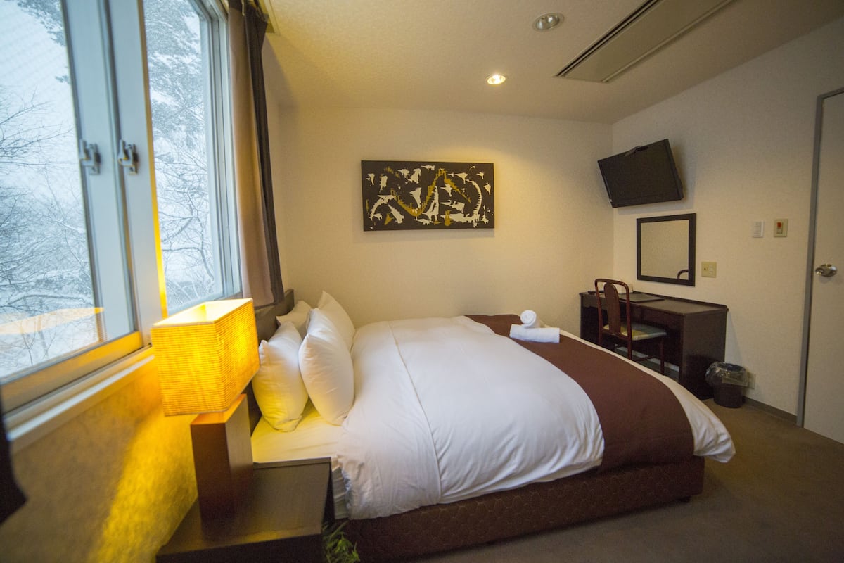 A hotel room with a bed, a desk, a chair, a TV, and a window with a view of trees.