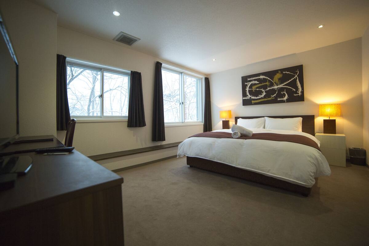 A hotel room with a large bed, two bedside lamps, a desk with a chair, and a window with curtains.