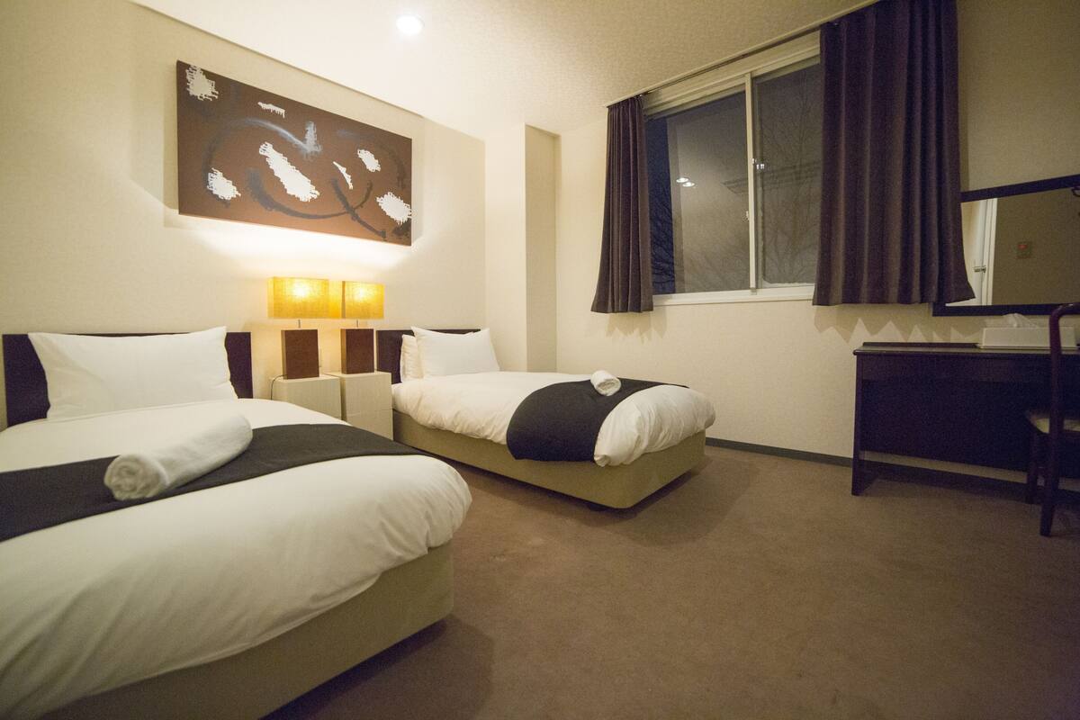 A hotel room with two beds, a desk, a window with curtains, and a wall-mounted artwork.