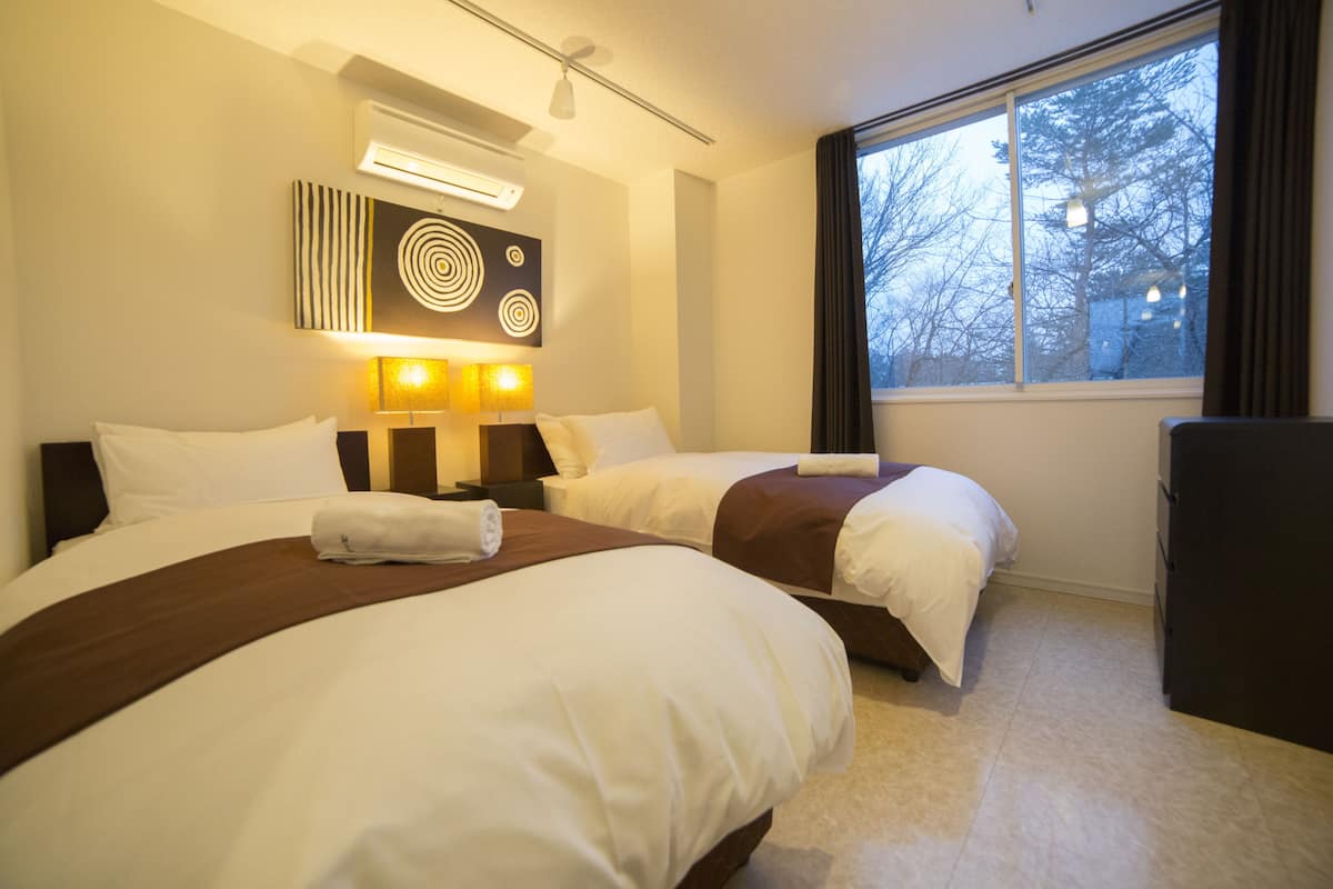 A hotel room with two beds, a large window with a view of trees, and a wall-mounted air conditioner.