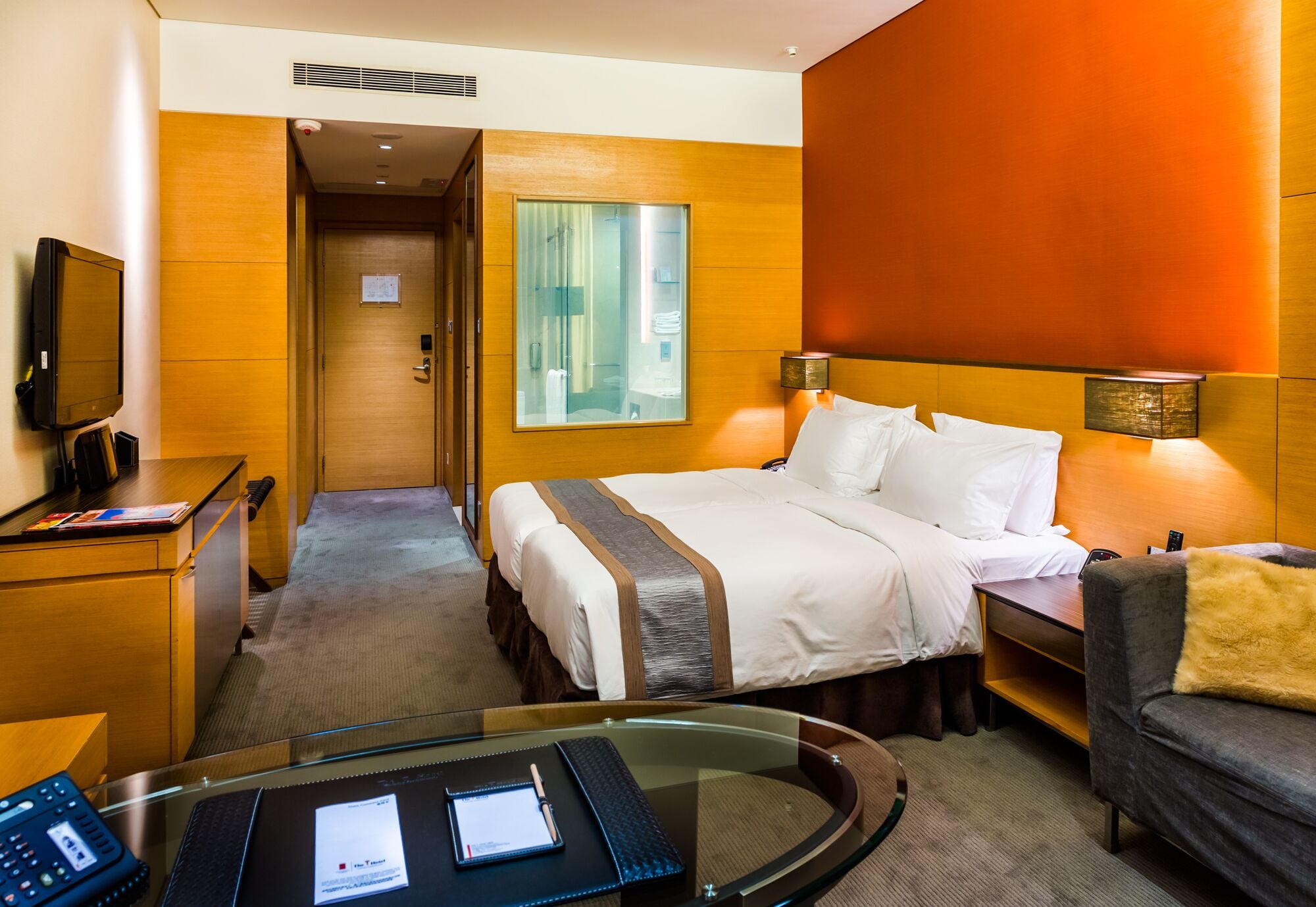 A hotel room with a bed, a desk with a computer, a television, and a glass table.