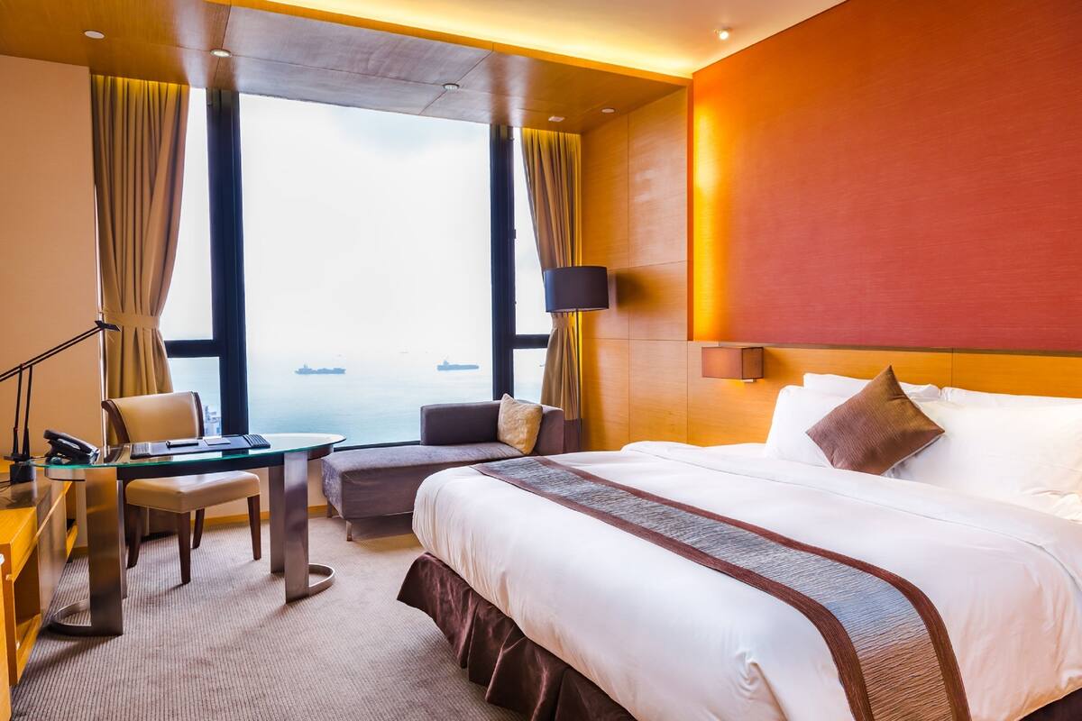 A hotel room with a large bed, a desk with a chair, a sofa, and a view of the sea through large windows.