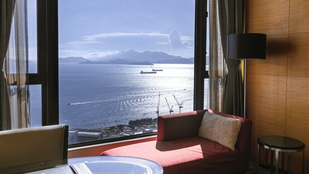 A room with a large window offering a view of the sea and mountains, a red sofa, a small round table, and a lamp. - The T Hotel (Hong Kong)