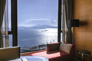 A room with a large window offering a view of the sea and mountains, a red sofa, a small round table, and a lamp. - The T Hotel (Hong Kong)