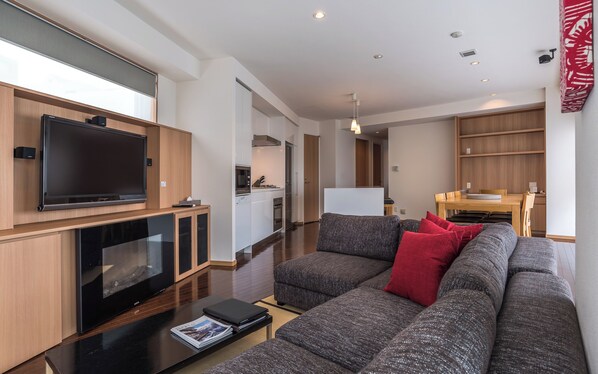Una sala de estar moderna con un televisor de pantalla plana grande, un sofá seccional gris con cojines rojos, una mesa de centro y una revista. Se observa un área de cocina al fondo con una mesa de comedor y sillas.
