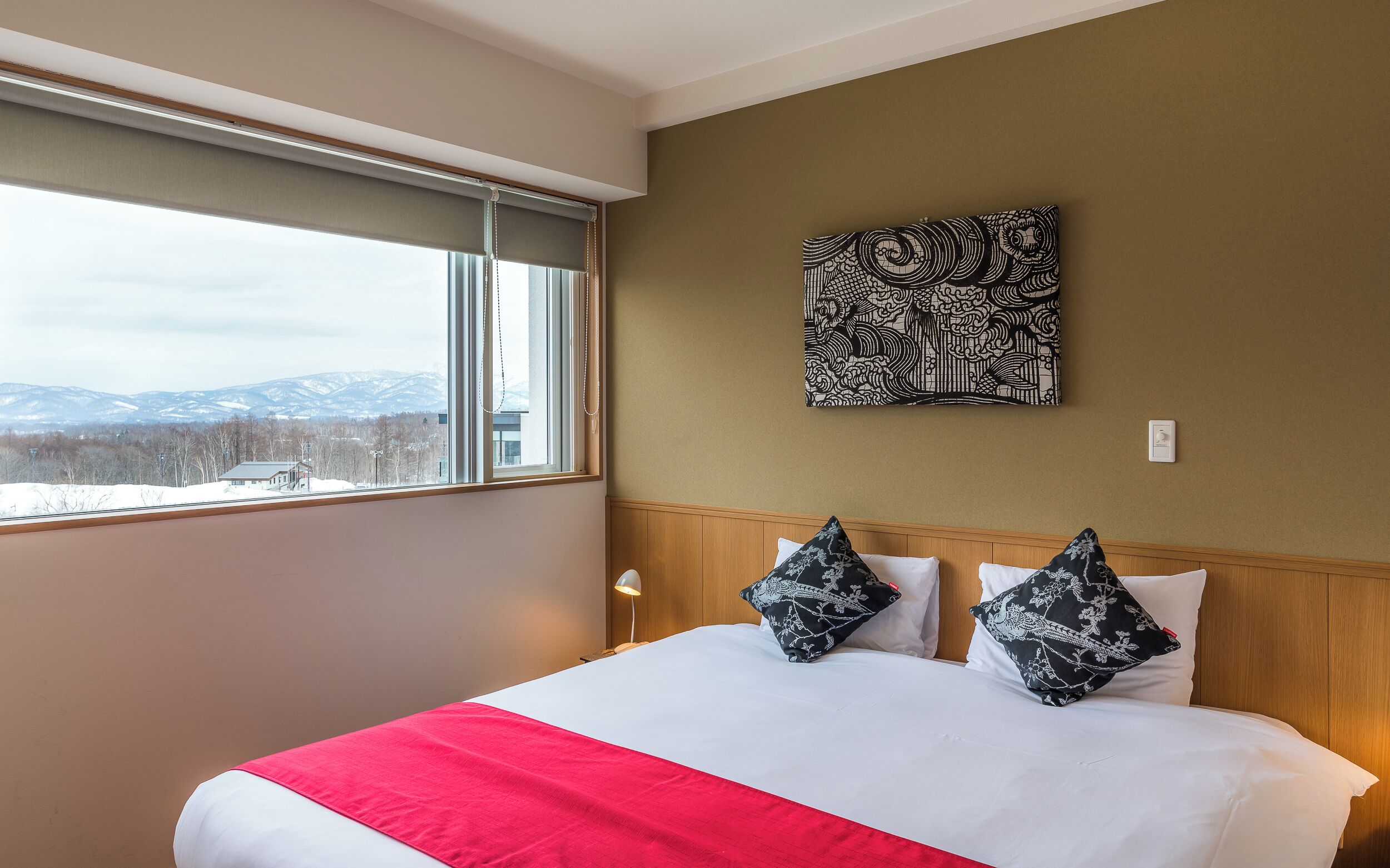 A hotel room with a large bed, two pillows, a red throw, a window with a view of mountains, and a wall-mounted artwork.