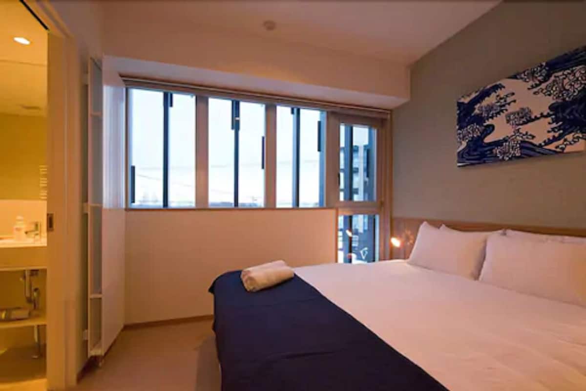 Ein Hotelzimmer mit einem großen Bett, einem Fenster mit Jalousien, einem Nachttisch und einem an der Wand hängenden Kunstwerk.