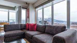 A modern living room with a large sectional sofa, a wooden coffee table, and large windows offering a view of snow-covered mountains. - Snow Crystal (Kutchan)