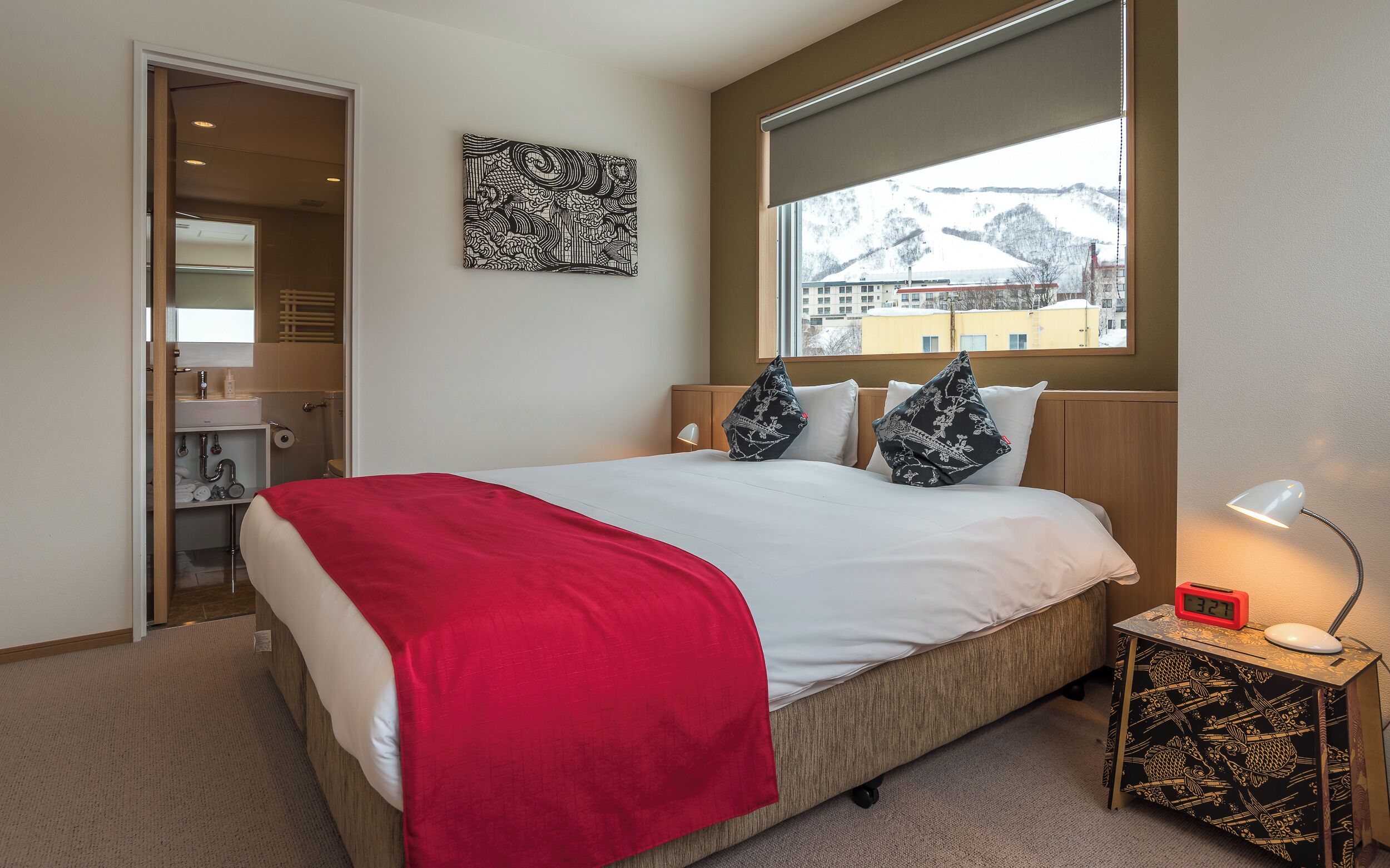 A hotel room with a bed, a bedside table with a lamp, a digital clock, and a window with a view of a snowy landscape.