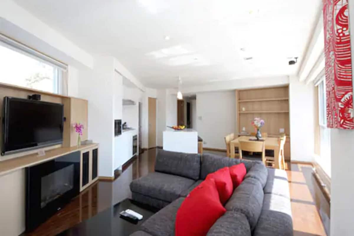 Ein modernes Wohnzimmer mit Flachbildfernseher, grauem Sofa mit roten Kissen, Holzboden und einem Essbereich mit Tisch und Stühlen.
