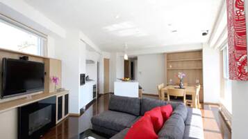 Una sala de estar moderna con un televisor de pantalla plana, sofá gris con cojines rojos, suelo de madera y una zona de comedor con mesa y sillas.