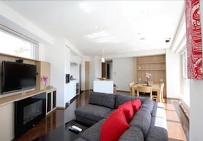 A modern living room with a flat-screen TV, grey sofa with red cushions, wooden flooring, and a dining area with a table and chairs. - Snow Crystal (Kutchan)