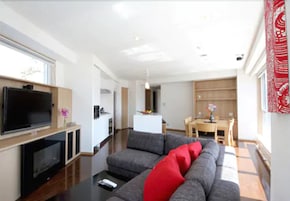 Una sala de estar moderna con un televisor de pantalla plana, sofá gris con cojines rojos, suelo de madera y una zona de comedor con mesa y sillas.