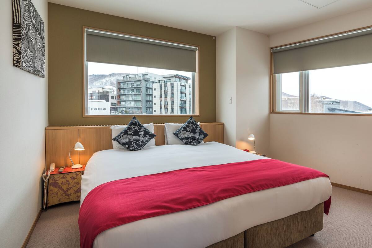 Ein Hotelzimmer mit einem großen Bett, zwei Nachttischlampen, einer roten Tagesdecke und Blick auf eine Stadtlandschaft durch die Fenster.