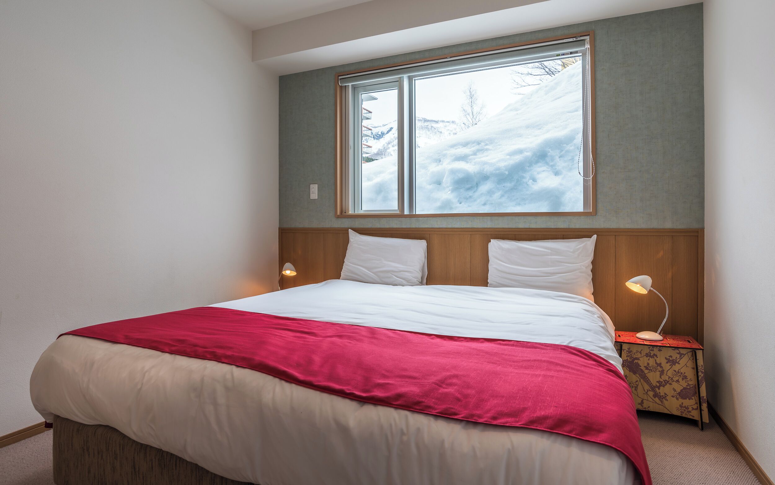 A bedroom with a large window, a bed with white and red bedding, and a nightstand with a lamp.