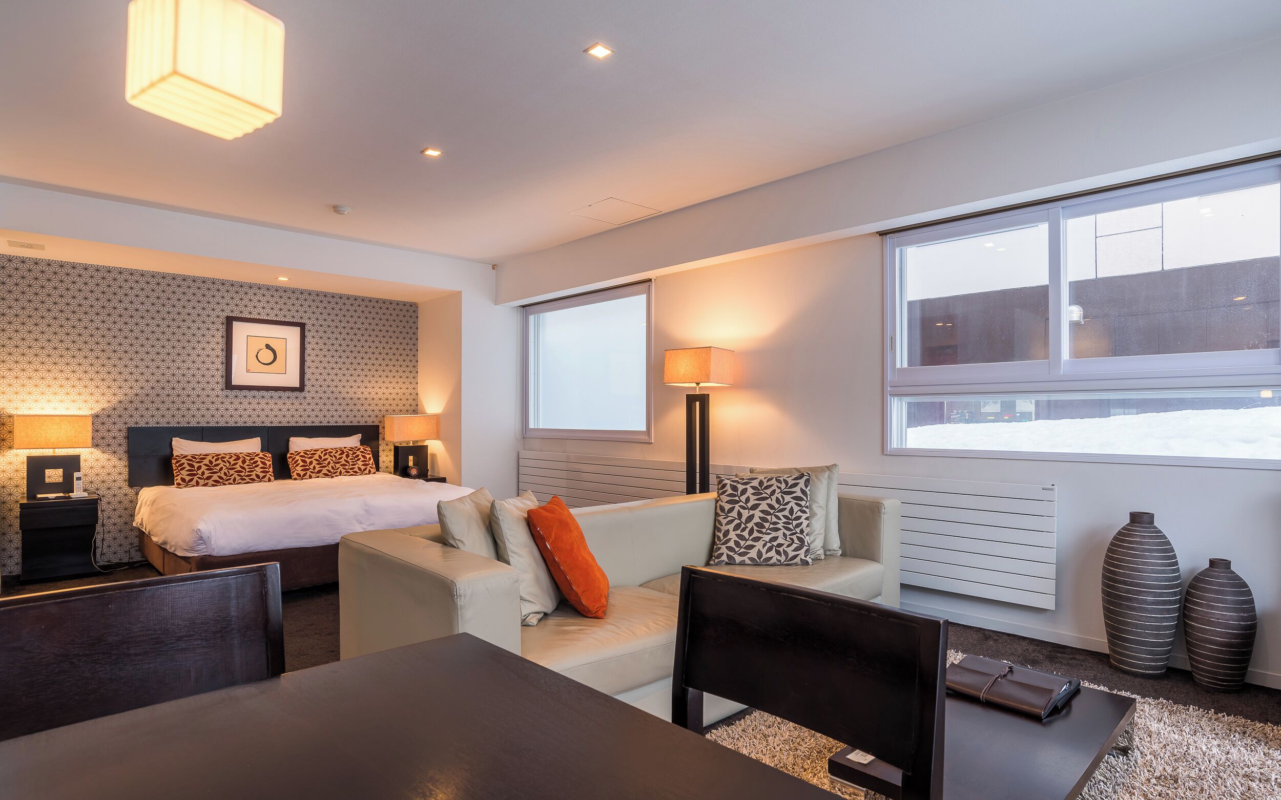 A modern hotel room with a bed, a sofa, a dining table, and a large window.