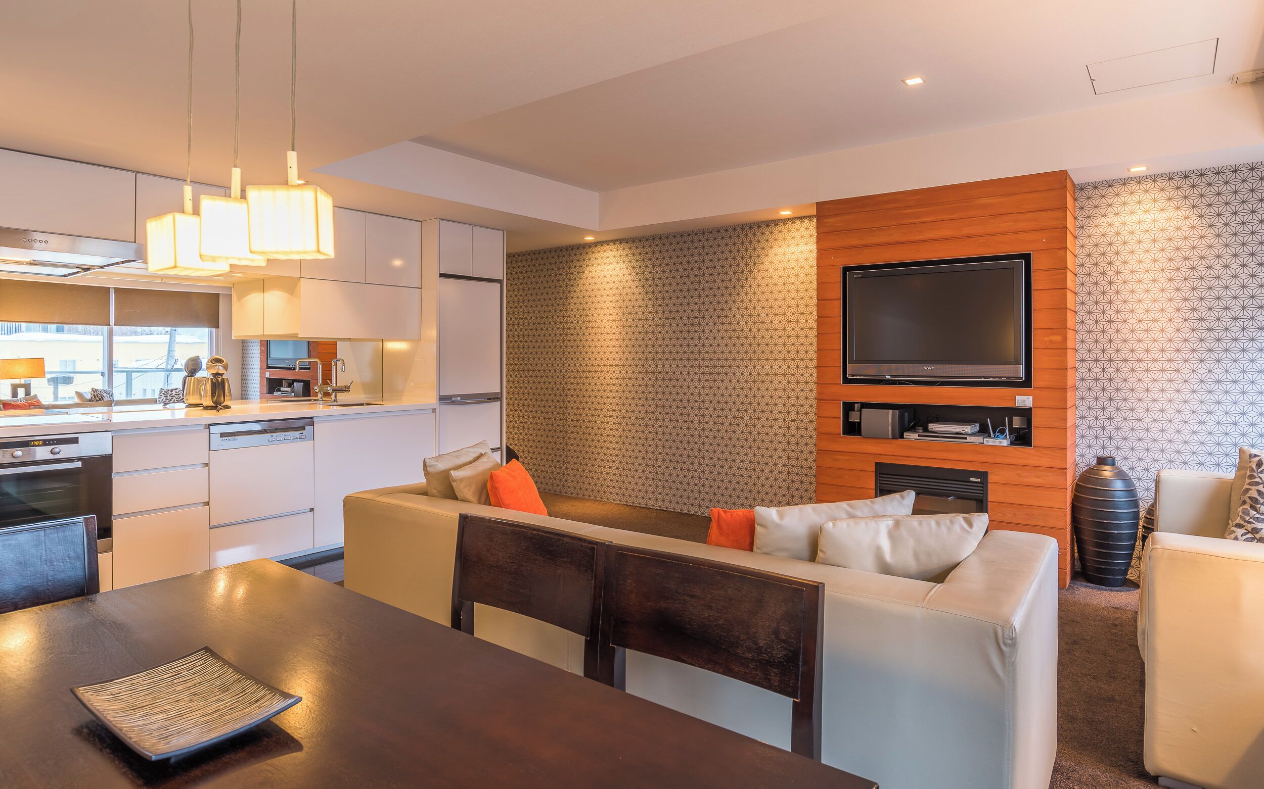 A modern kitchen with built-in appliances, a dining area with a dark table, and a living area with a sofa and a TV mounted on a wooden wall.