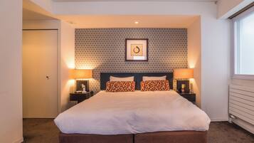 A neatly arranged hotel room with a large bed, two bedside tables with lamps, a patterned wallpaper, and a framed picture on the wall.