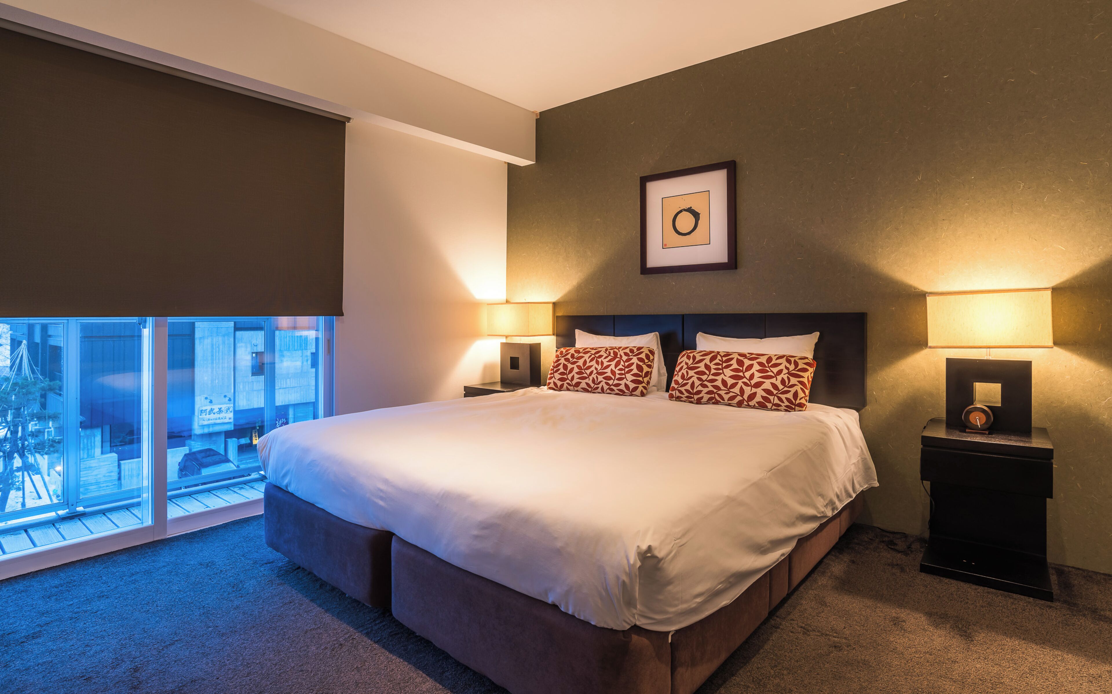 A bedroom with a large bed, two bedside tables with lamps, a framed picture on the wall, and a view through a window.