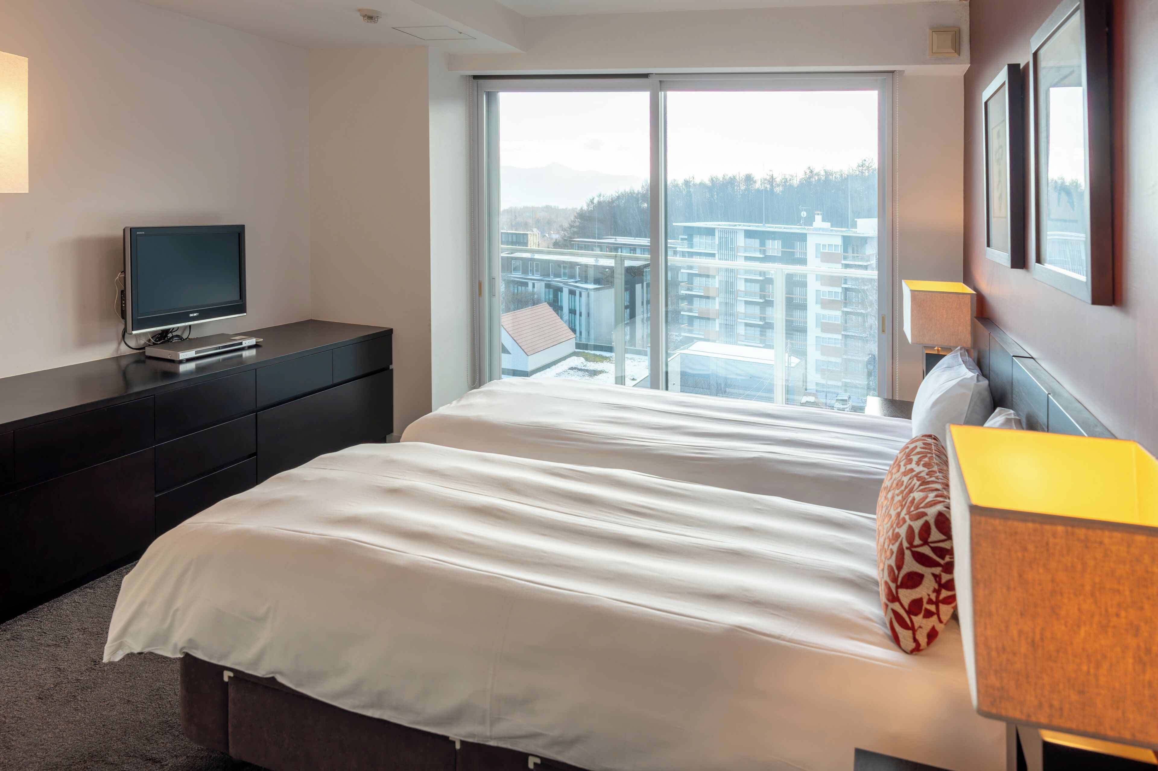 Una habitación de hotel moderna con una cama grande, un televisor de pantalla plana, un escritorio con lámpara y vistas a la ciudad a través de amplios ventanales.