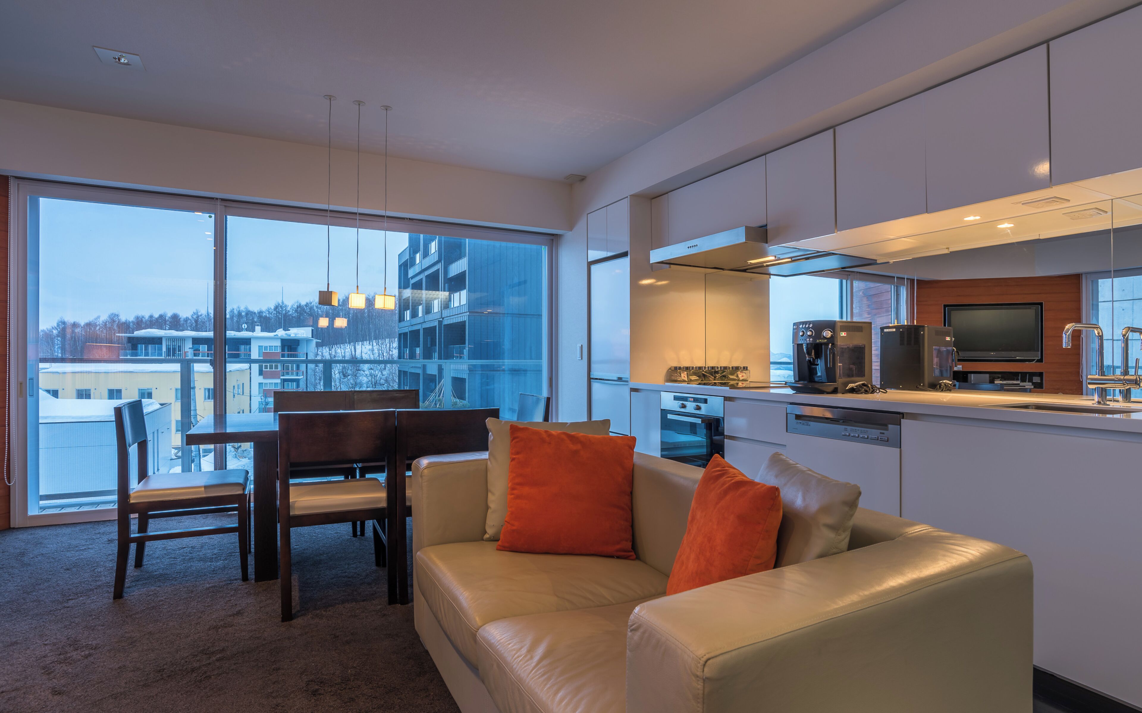 A modern apartment with a dining area, a sofa with orange cushions, a kitchen with appliances, and large windows offering a city view.