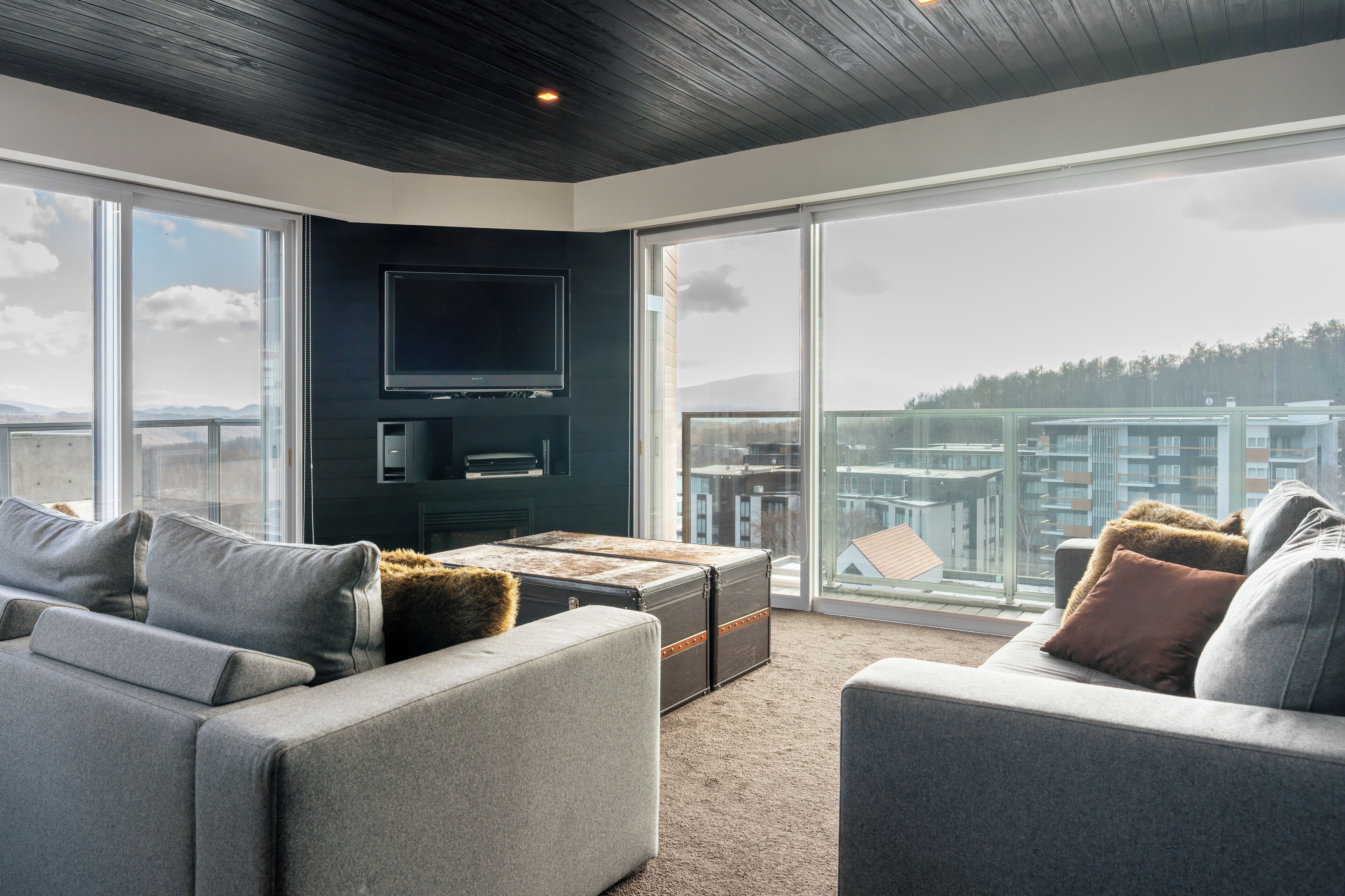 A modern living room with a large flat-screen TV, grey sofas, and a view of a cityscape through large windows.