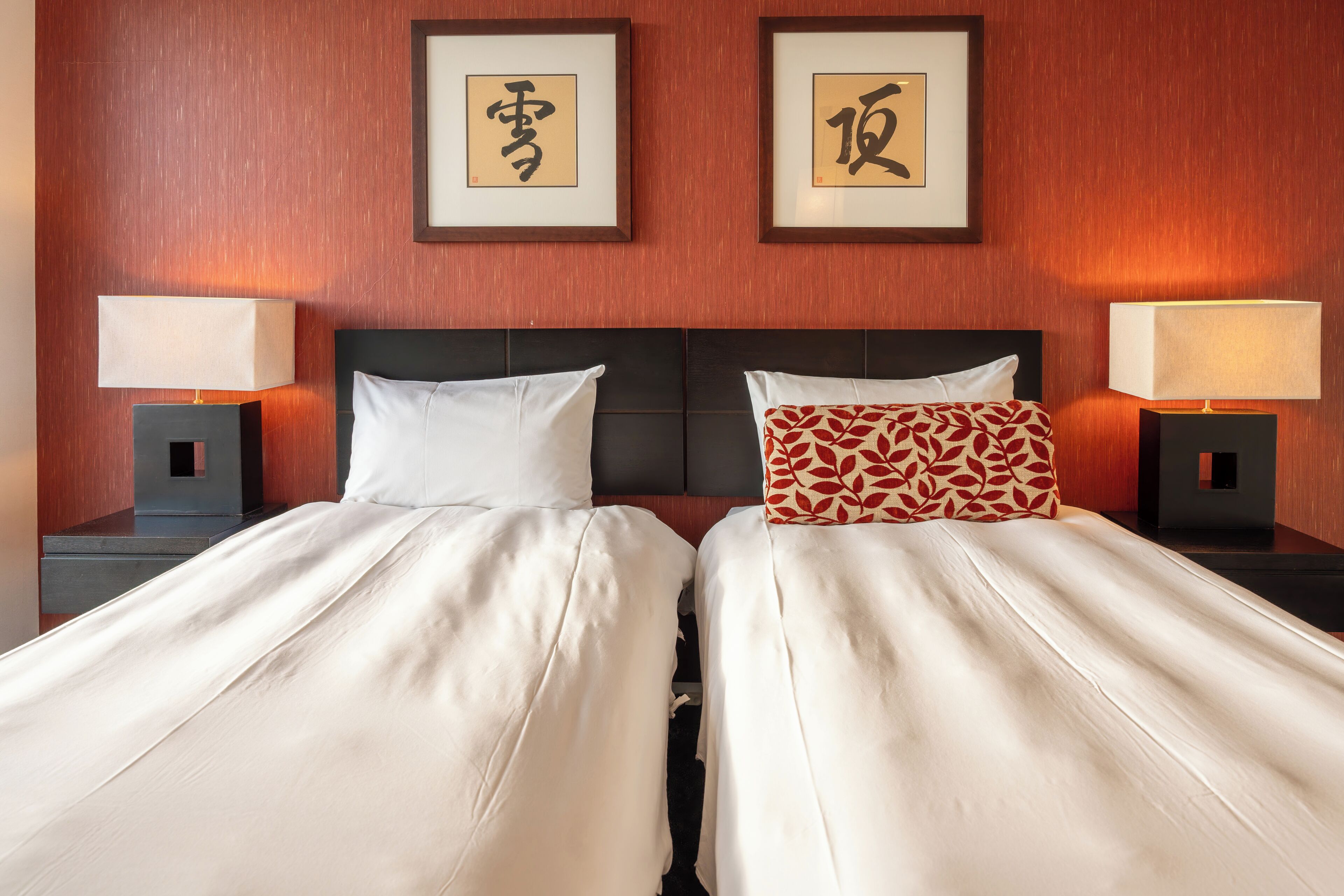Ein Hotelzimmer mit zwei Betten, jeweils mit weißer Bettdecke und Kissen. Über den Betten hängen gerahmte Kalligraphie-Werke an der Wand.