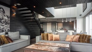 Una sala moderna con un sofá gris, un gran baúl marrón y una escalera que lleva a un nivel superior.