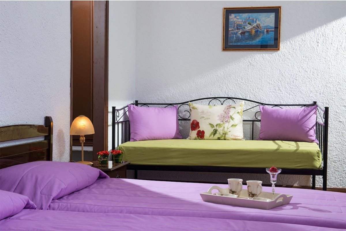 Ein Bett mit einer grünen Tagesdecke und zwei violetten Kissen, ein Nachttisch mit einer Lampe sowie ein Tablett mit Tassen und einem Dessert.
