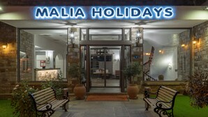 The entrance to Malia Holidays with a stone facade, two benches, and a clear view into the lobby.