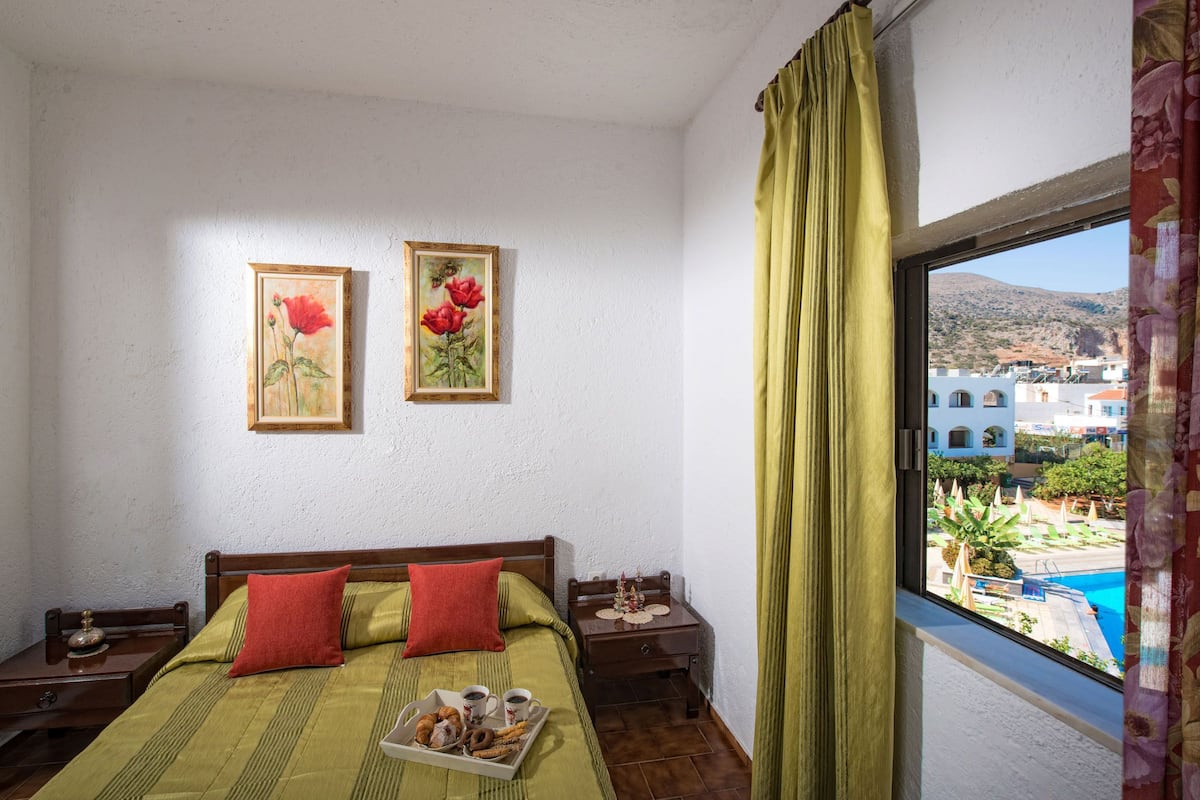 Ein Schlafzimmer mit einem Bett, zwei gerahmten Blumenbildern, einem Nachttisch mit einer Snackauswahl und einem Fenster mit Blick auf Gebäude und einen Pool.