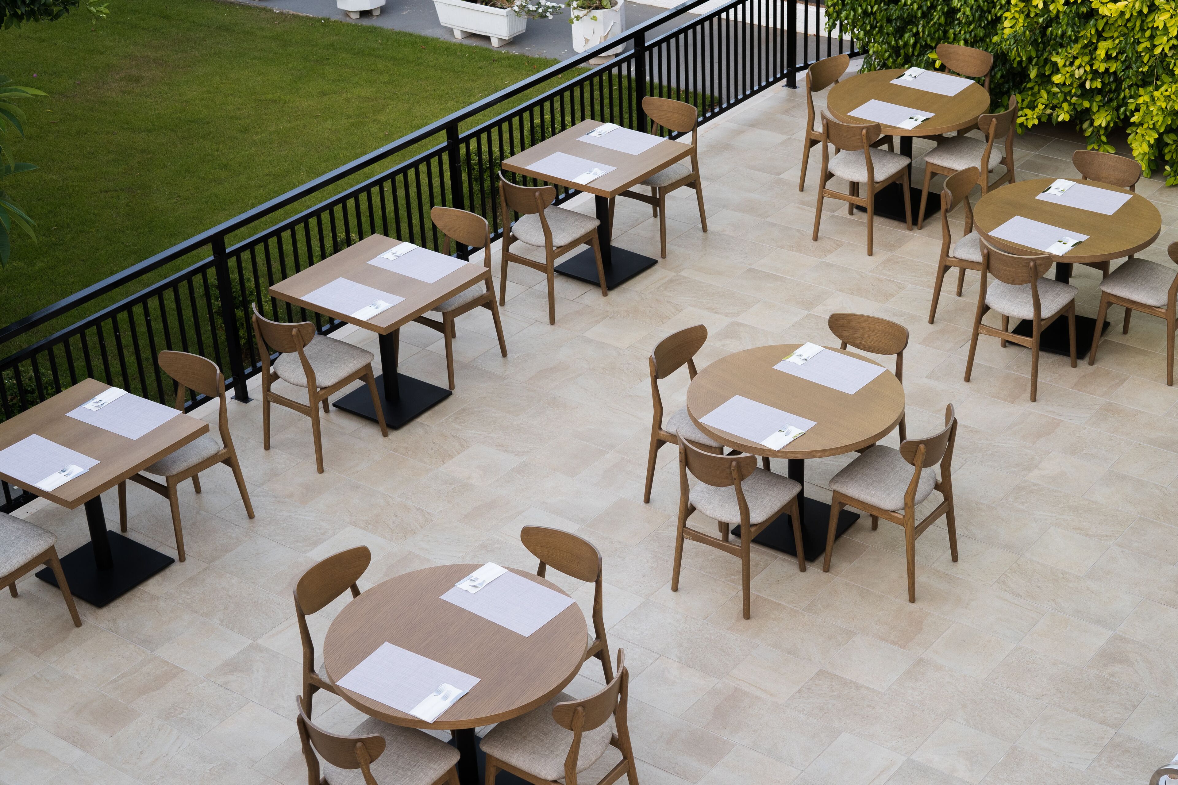 Een terras buiten met verschillende ronde en rechthoekige tafels en stoelen, geplaatst op een betegelde vloer met een zwarte balustrade.