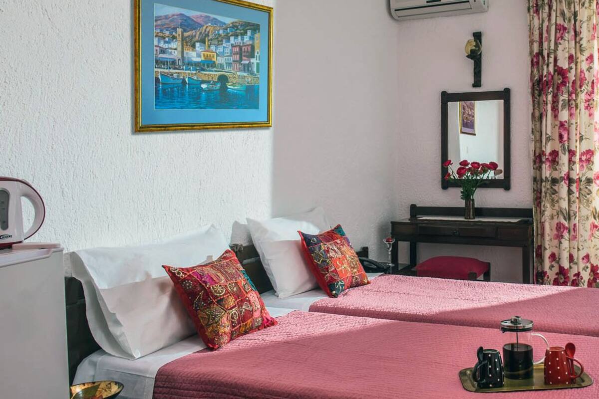 Ein Hotelzimmer mit zwei Betten, einem Schreibtisch, einem Spiegel, einem Gemälde und einem blumigen Vorhang.