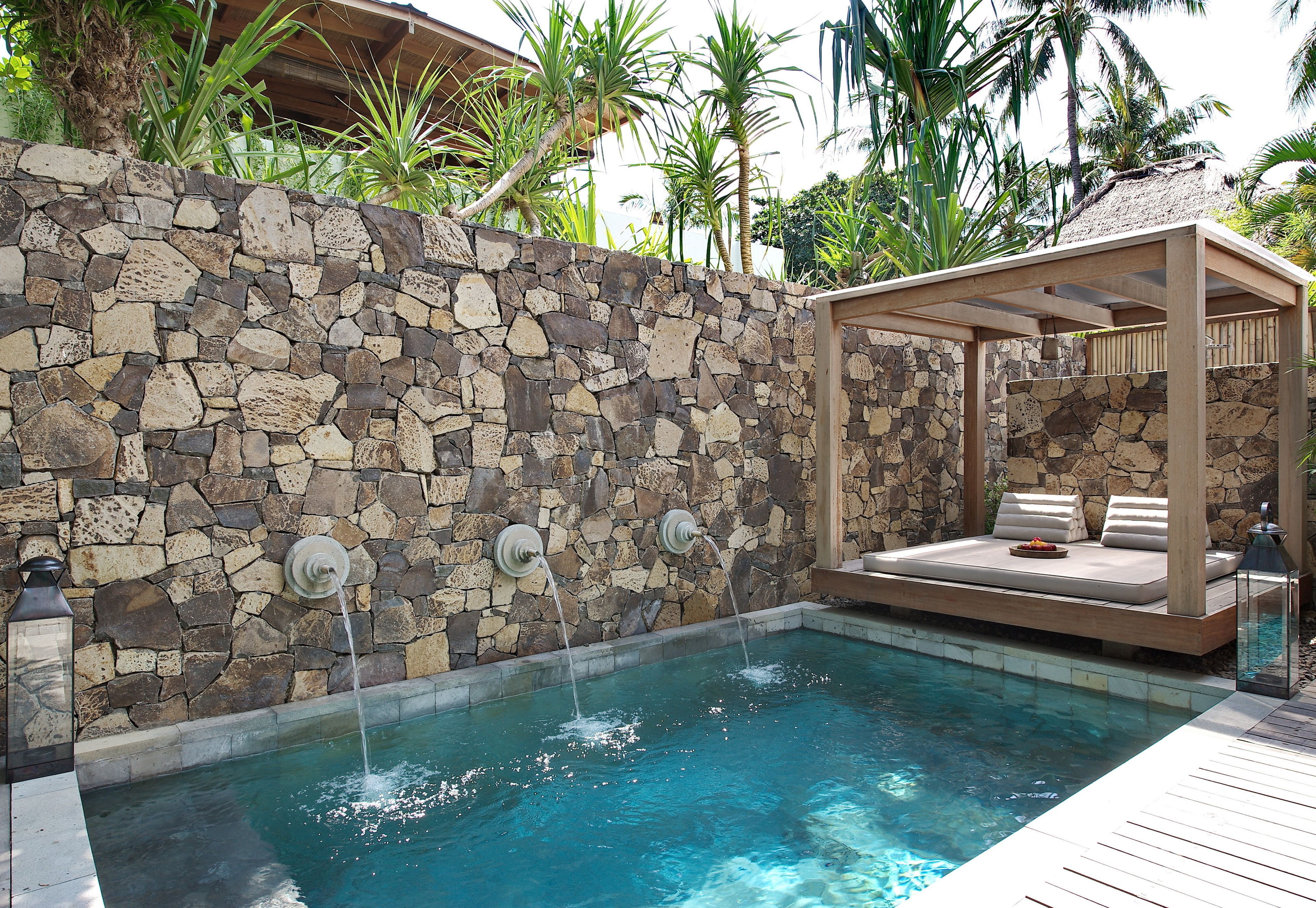 Eine Steinmauer mit Pool, eine Holzterrasse und eine überdachte Sitzgruppe mit Kissen.