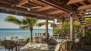 An outdoor dining area with wooden tables and chairs, a palm tree, and a view of the ocean. - Jeeva Santai Villas (Senggigi)