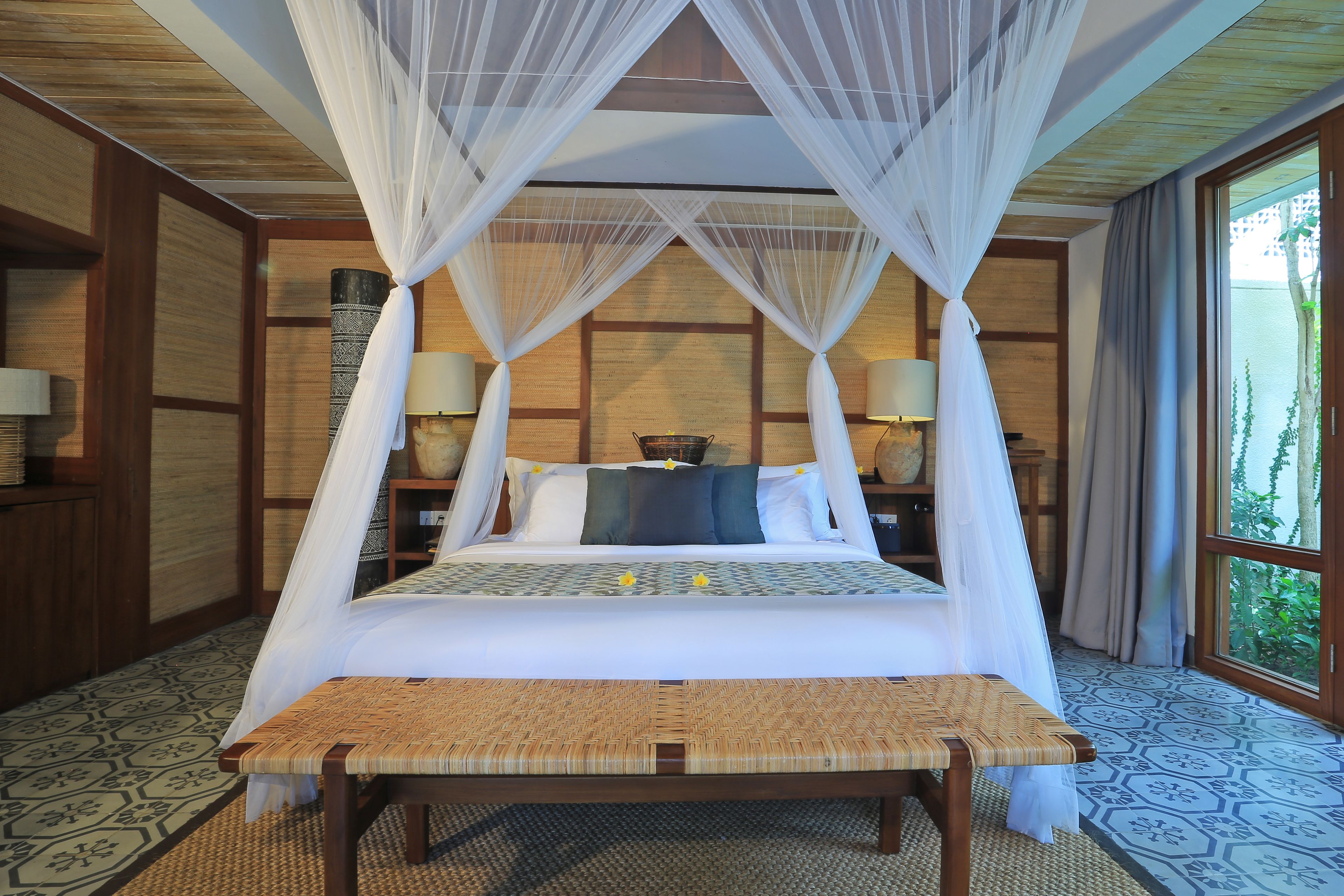 A four-poster canopy bed with a woven bench in front, a nightstand with lamps, and a view of greenery through a large window.