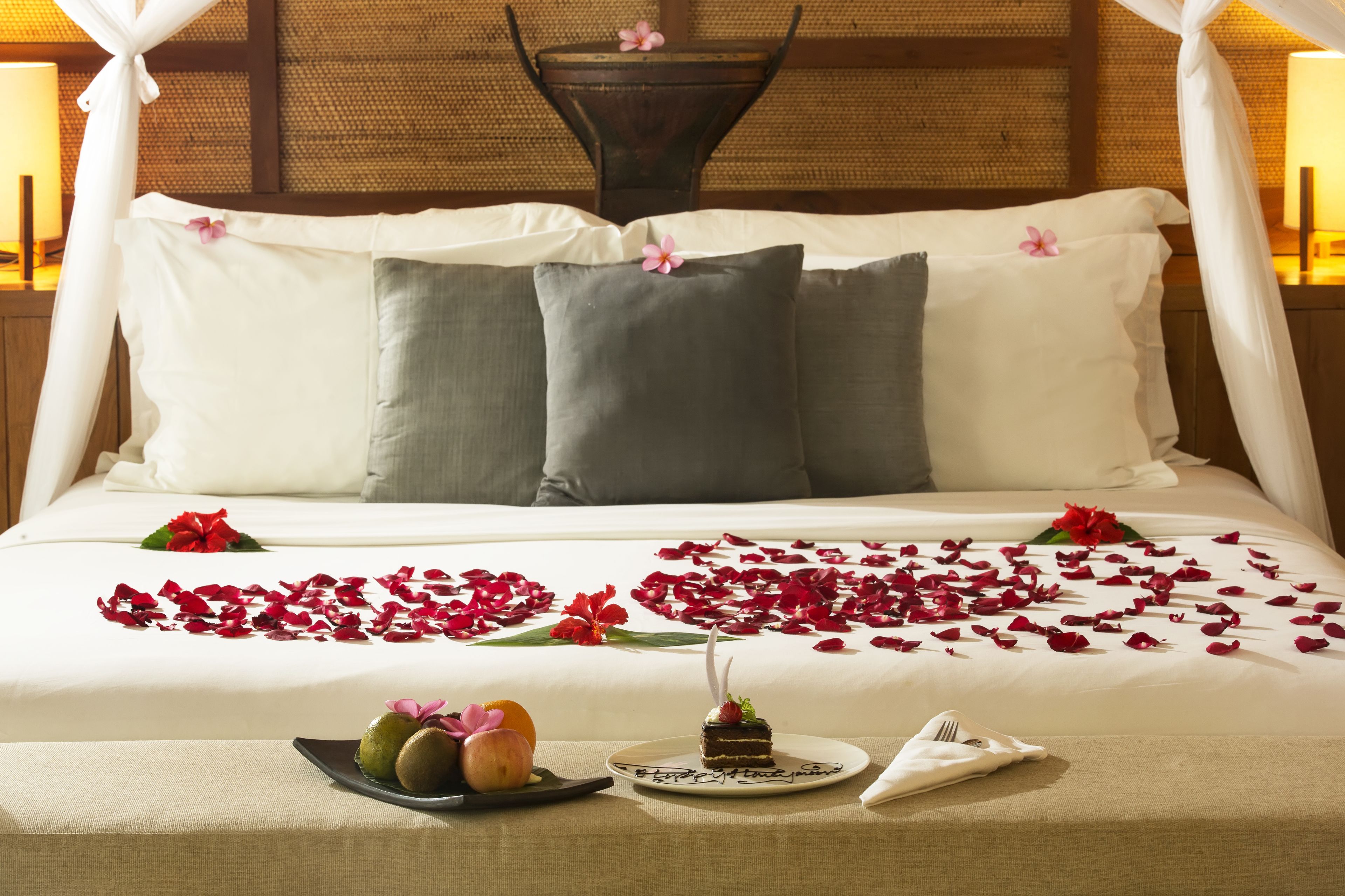 Una cama elegantemente arreglada con dosel, decorada con pétalos de rosa y un pequeño arreglo floral.