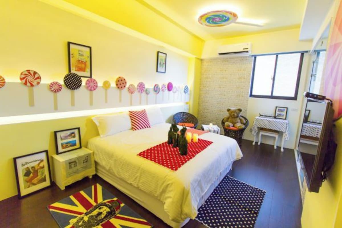 A bedroom with a bed, bedside tables, a teddy bear, a small table with a lamp, and a window.