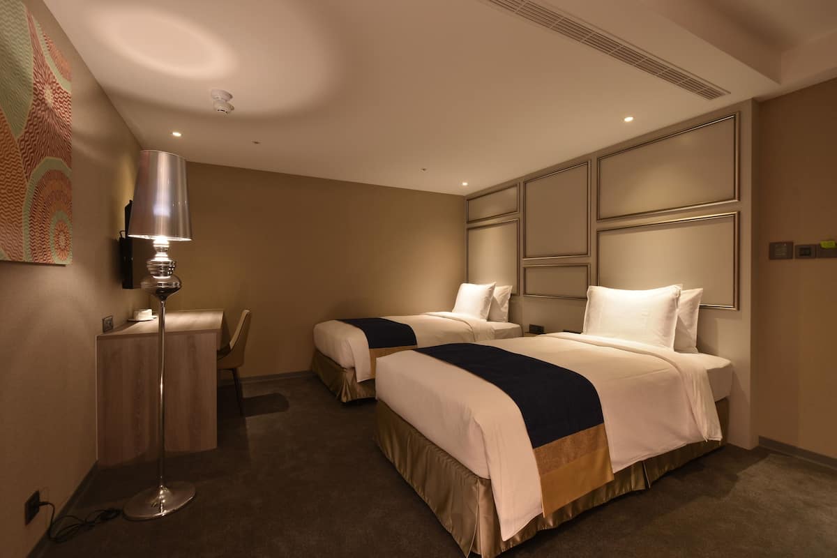 Habitación de hotel con dos camas, un escritorio, una lámpara y una obra de arte en la pared.