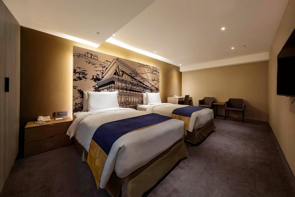 Una habitación de hotel con dos camas, un escritorio pequeño, una silla y un televisor en la pared.